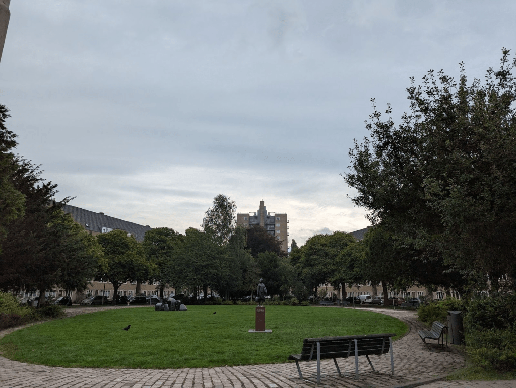 Picture taken Aug 03, 2025 of park at Merwedeplein. The skyscraper can still be seen in the background. In the middle ground is a statue of Anne Frank. It's hard to see the apartment buildings on Merwedeplein because of the trees.